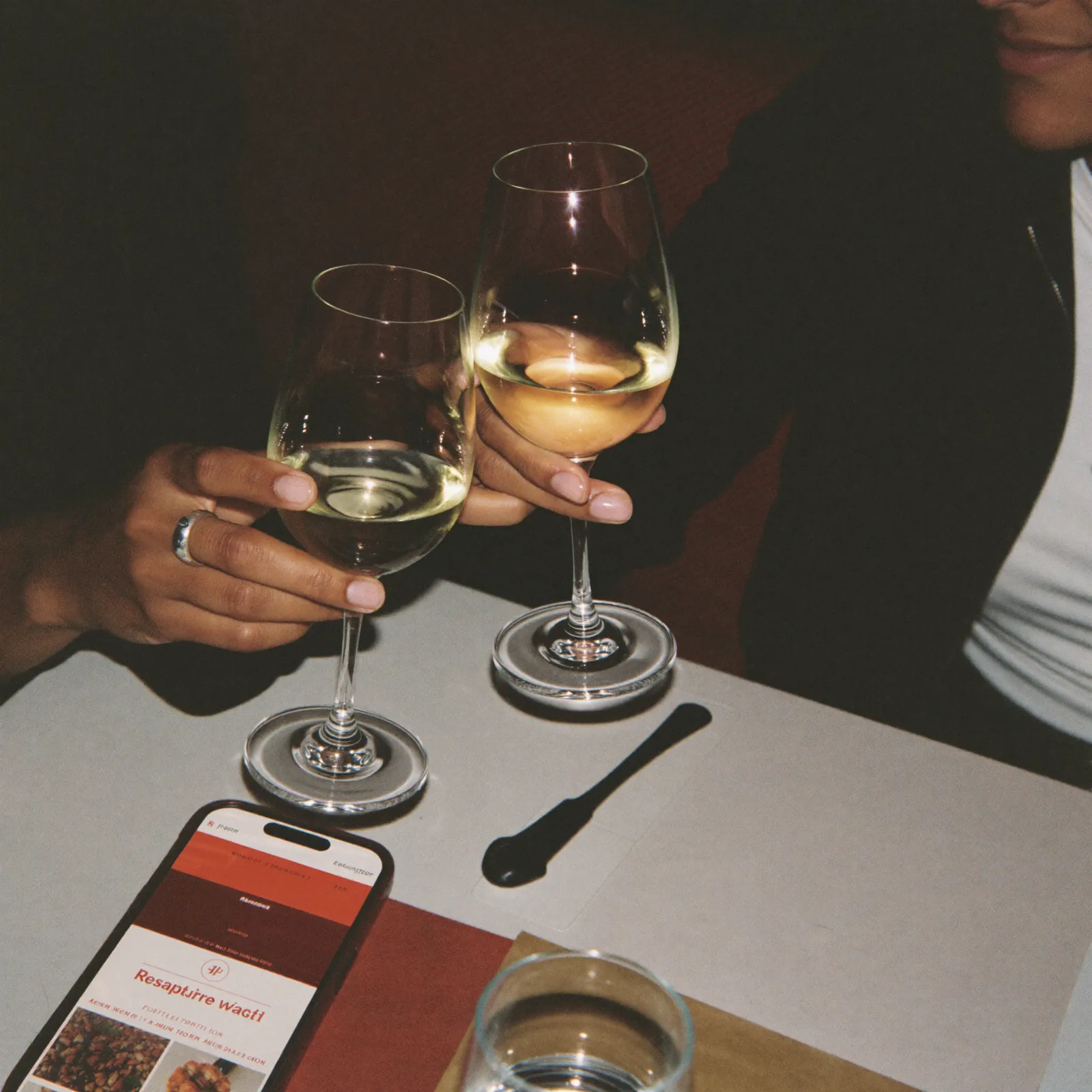 Two friends clinking wine glasses at a restaurant table with a phone showing the restaurant's website